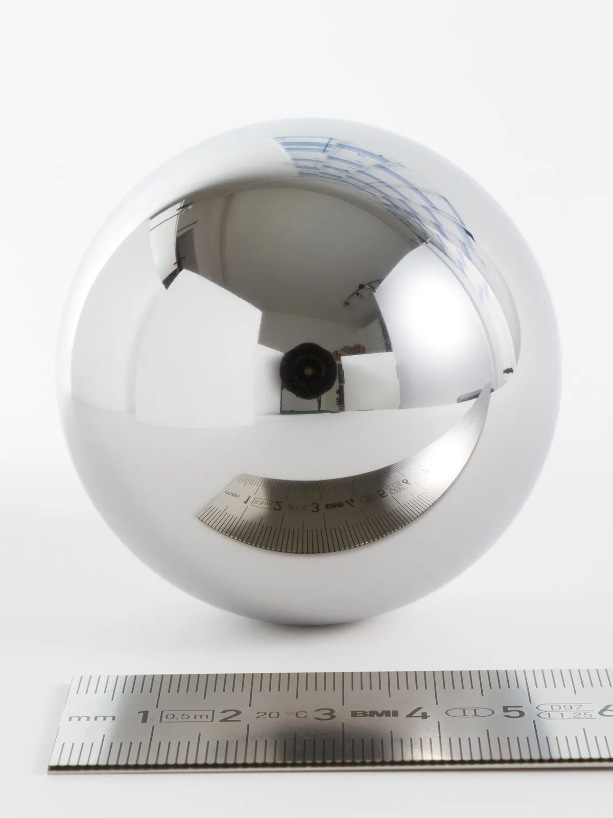 Esfera de aço polido, de uns seis centímetros, sobre fundo branco ao lado de uma régua de metal graduada em milímetros; a esfera reflete o ambiente como um espelho.