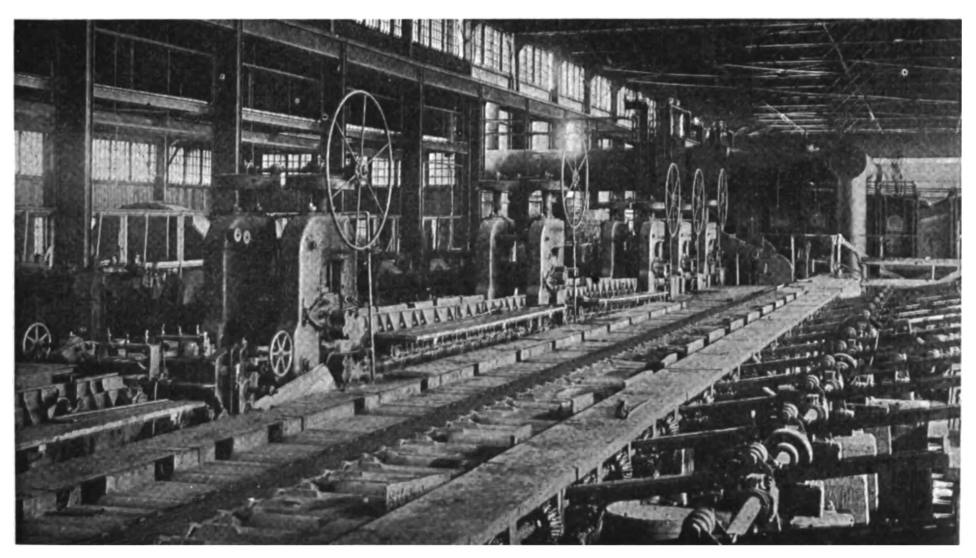 Fotografia em preto e branco de 1919: o interior de uma laminação de aço, com uma longa mesa de rolos ao centro e grandes volantes de ferro nas máquinas ao fundo, sob um telhado de vidro.