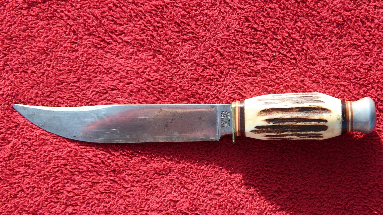 Faca de caça com lâmina larga marcada "Baron Solingen Germany", cabo de chifre de veado com a superfície rugosa em tons de creme e marrom, virola e pomo de metal, sobre um pano vermelho.