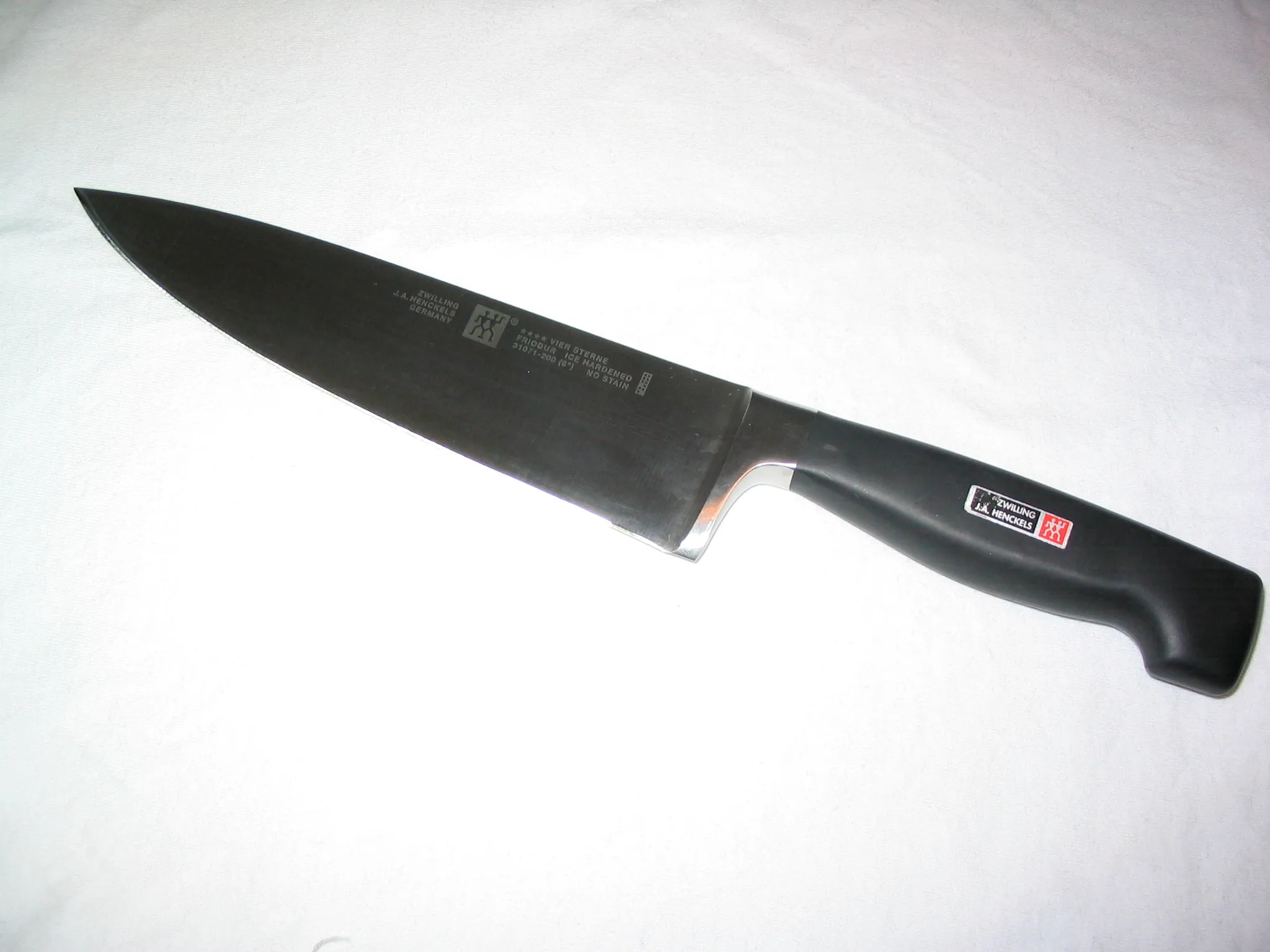 Faca de chef Zwilling de oito polegadas sobre um pano branco, com a lâmina escura de reflexo, o talão de aço polido entre a lâmina e o cabo preto, e a marca dos gêmeos gravada.