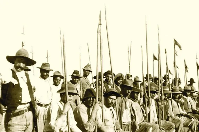 Fotografia em sépia de 1923: dezenas de homens de chapéu e lenço, sentados e em pé, com lanças compridas erguidas; um deles, em primeiro plano, à esquerda, de bigode, lenço claro e cinturão com faca.