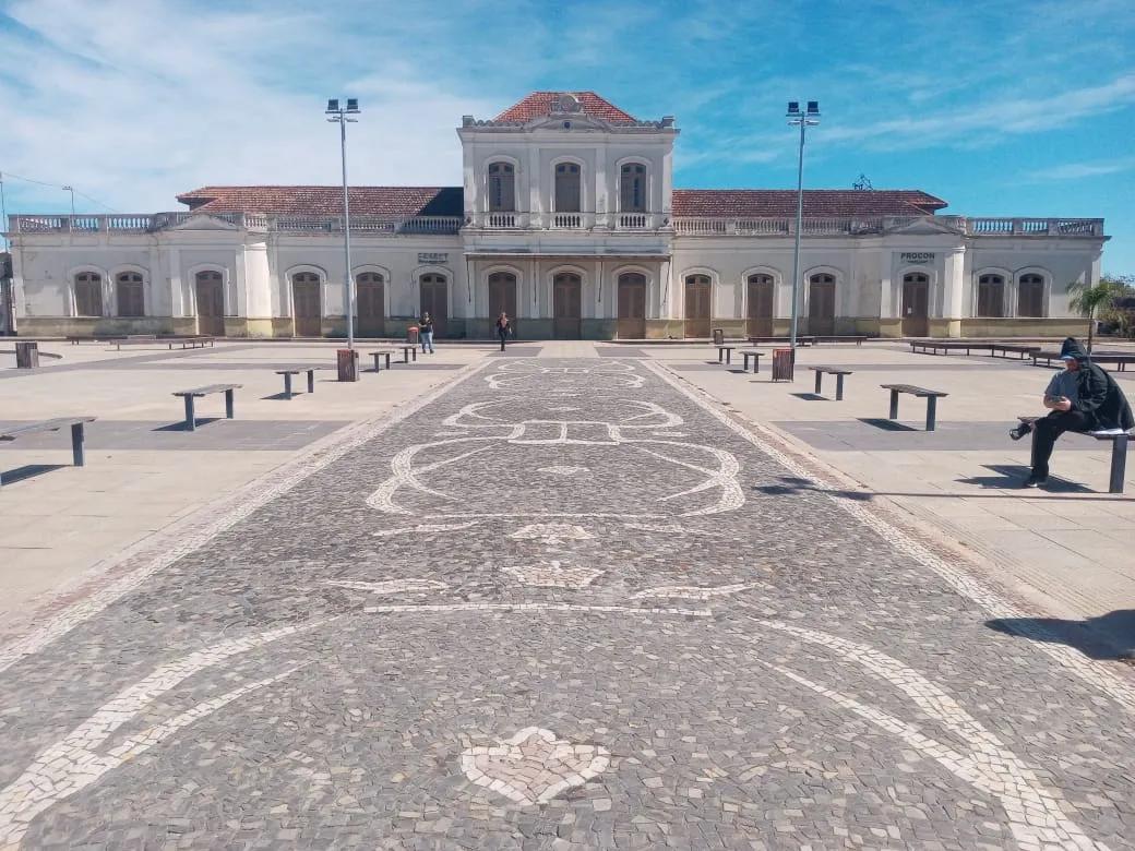 Fachada de estação ferroviária de estilo eclético, com torre central, telhado de telhas e pátio de pedras portuguesas em desenho geométrico, sob céu azul, em Pelotas