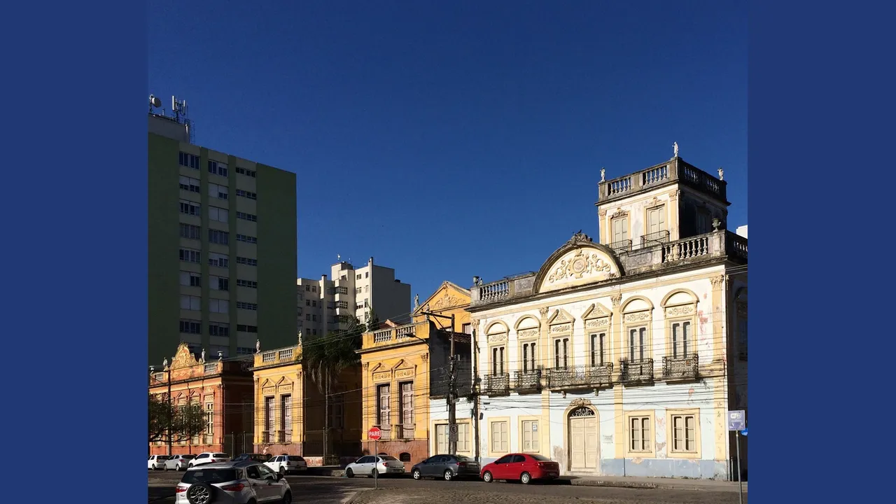 Fileira de casarões históricos de fachada eclética, em tons de amarelo, branco e vermelho, com sacadas de ferro trabalhado e platibandas ornamentadas, numa praça de pedras portuguesas de Pelotas, sob céu azul.