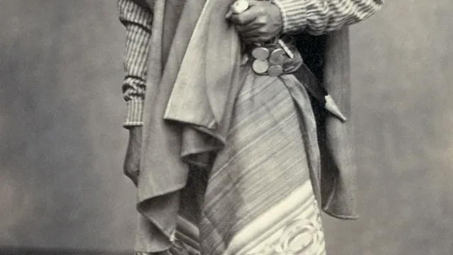Retrato de estúdio de um gaúcho barbado de 1868, com chapéu, poncho e chiripá, tirador com rastra de moedas de prata na cintura e o cabo de um facão à mostra.