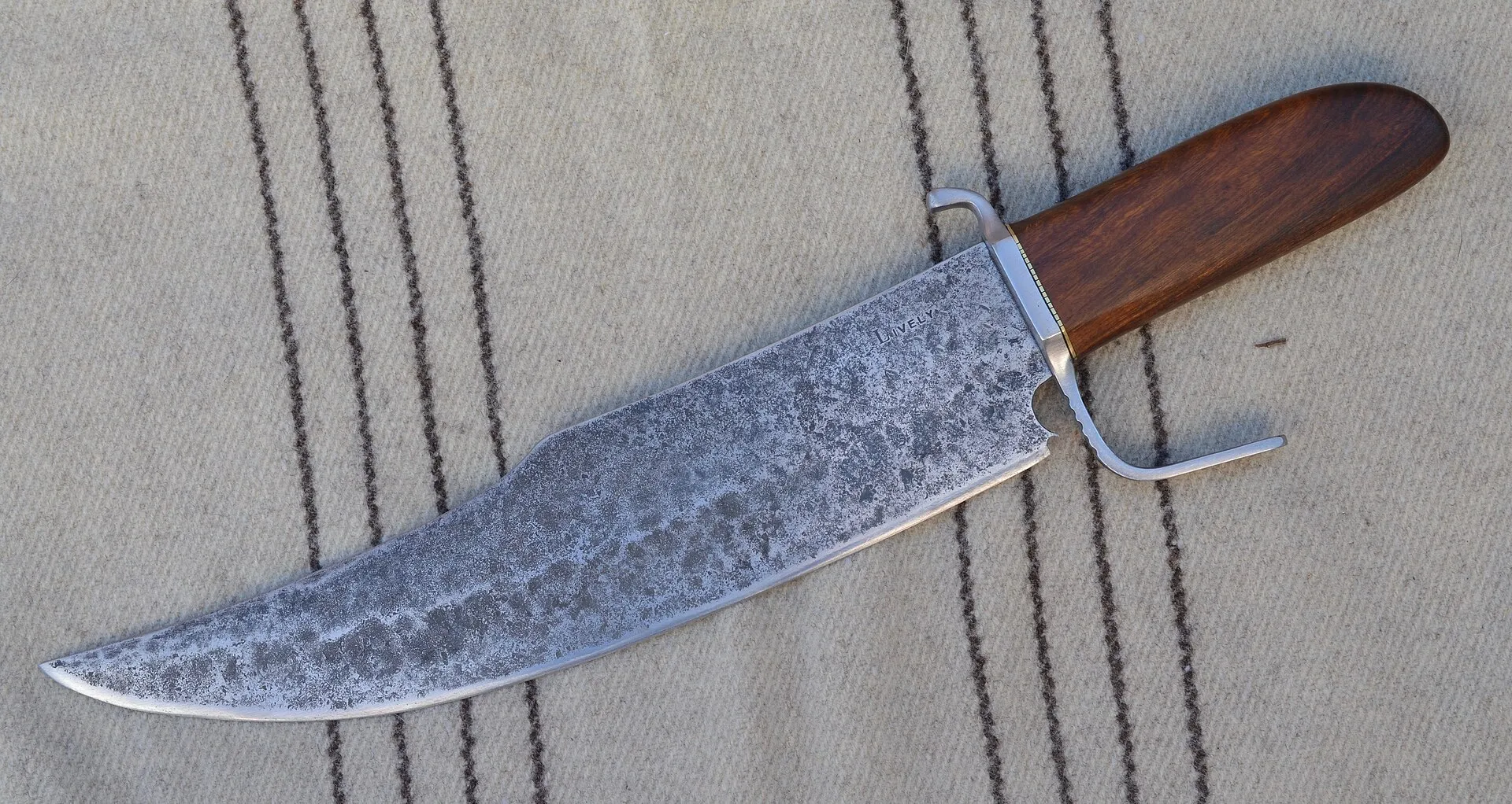 Faca bowie forjada à mão, lâmina larga com marcas escuras de forja e fio polido, guarda de aço e cabo de madeira escura, apoiada sobre um cobertor bege