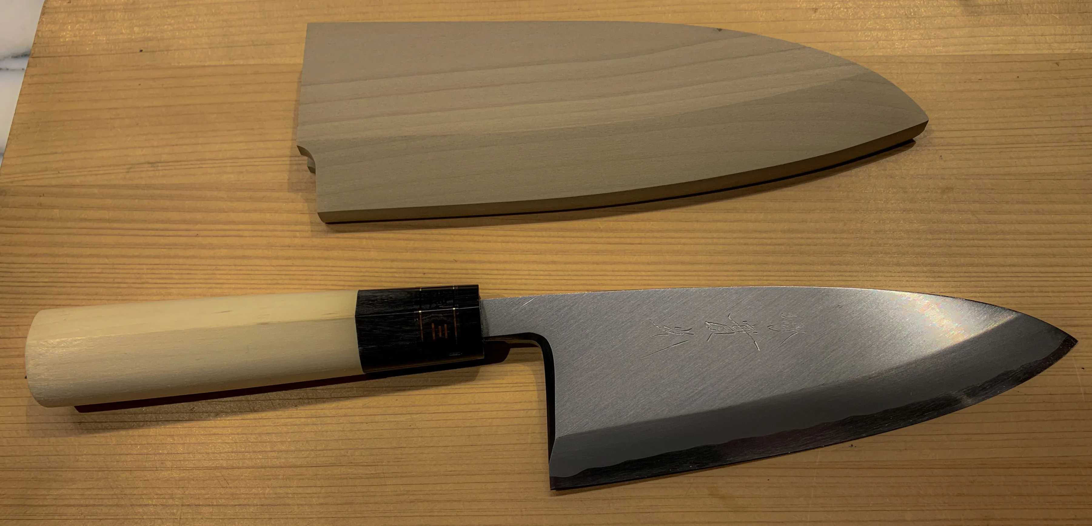 Faca japonesa de cozinha do tipo deba sobre uma tábua de madeira clara, com cabo de magnólia e virola escura, a lâmina larga com inscrição gravada em japonês; acima dela, a bainha de madeira.