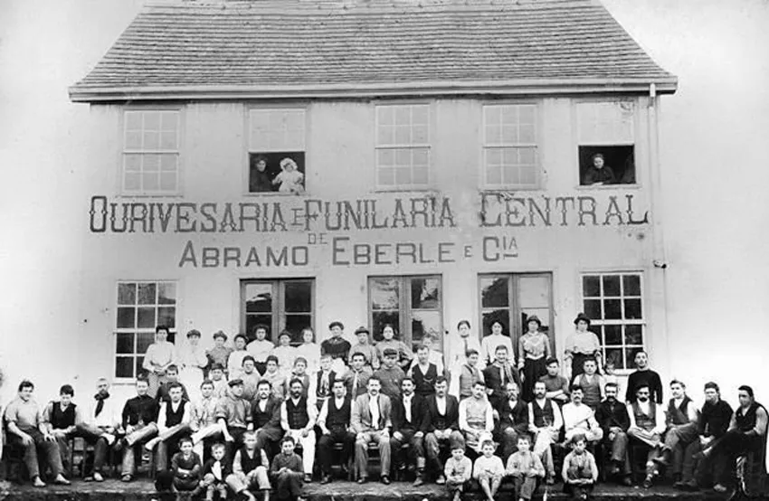 Fachada de sobrado com os dizeres "Ourivesaria e Funilaria Central — Abramo Eberle e Cia", com dezenas de operários, mulheres e crianças posando em frente, em 1907.