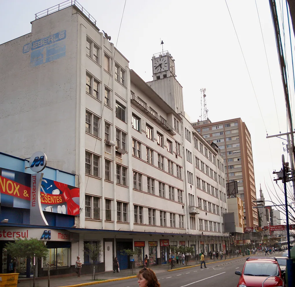 Prédio industrial branco de cinco andares com torre de relógio, o nome EBERLE ainda pintado na empena lateral, com lojas no térreo e movimento de rua