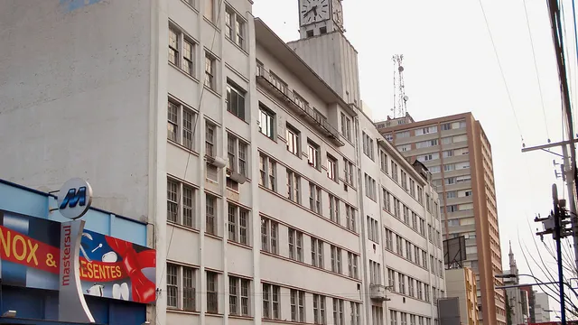 Prédio industrial branco de cinco andares com torre de relógio, o nome EBERLE ainda pintado na empena lateral, com lojas no térreo e movimento de rua, em Caxias do Sul.