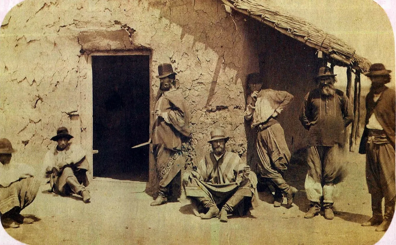 Grupo de gaúchos em frente a um rancho de adobe com teto de palha, em fotografia sépia da década de 1860; o homem de poncho em pé, ao centro, segura um facón de lâmina nua
