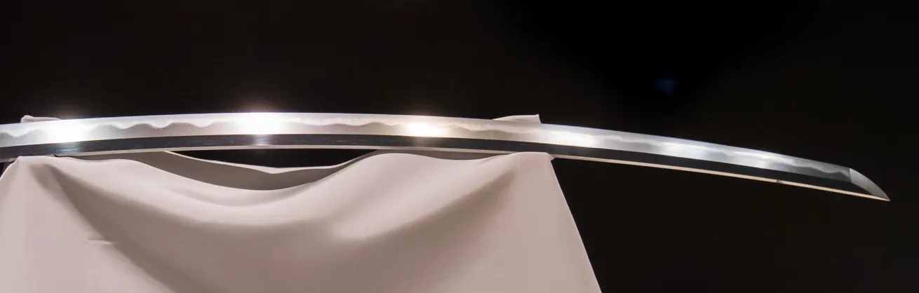 Close de uma lâmina de katana sobre um apoio de tecido branco, com o fundo escuro; ao longo do fio, uma faixa ondulada mais clara se destaca do resto do aço.