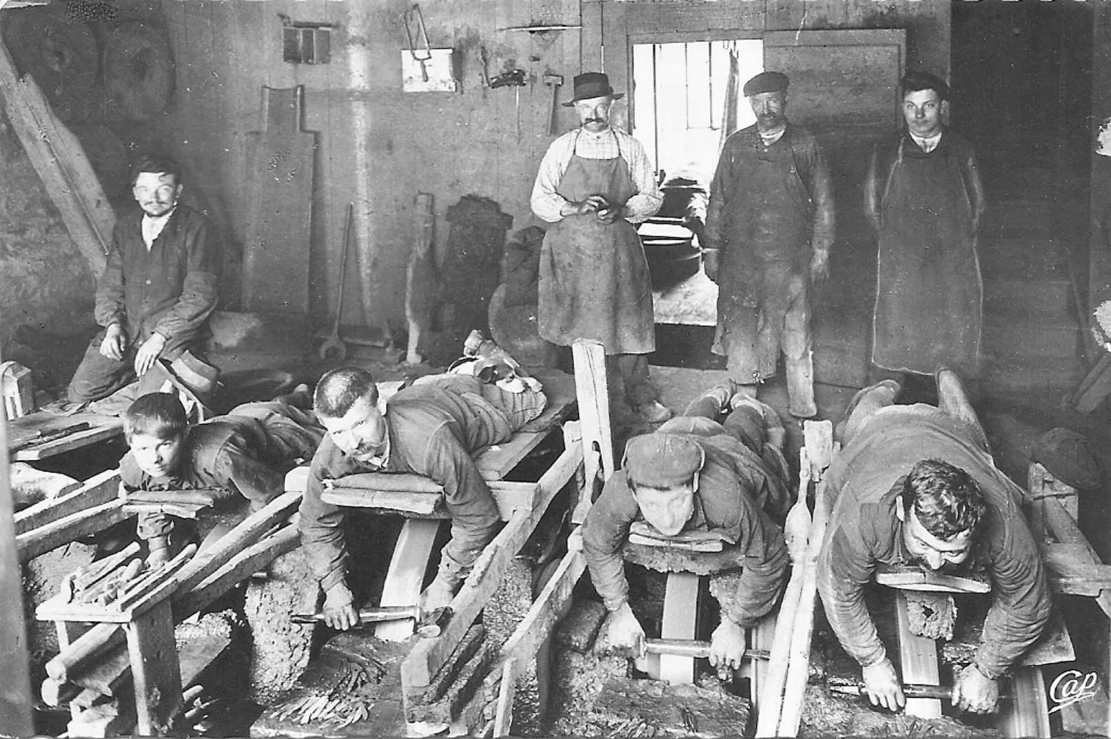 Fotografia antiga de uma oficina de cutelaria em Thiers, com cinco homens deitados de bruços sobre pranchas de madeira, debruçados sobre rebolos de pedra girando na água, enquanto três outros posam de pé ao fundo