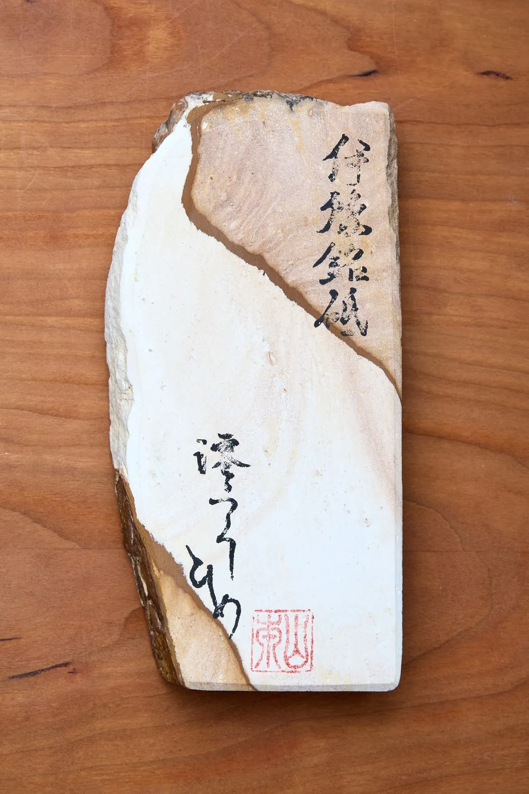 Pedra de afiar natural japonesa, em tons de bege e branco, vista de cima sobre uma mesa de madeira; a face tem uma camada clara e outra mais escura, e traz inscrições em japonês a pincel preto e um carimbo vermelho no canto.