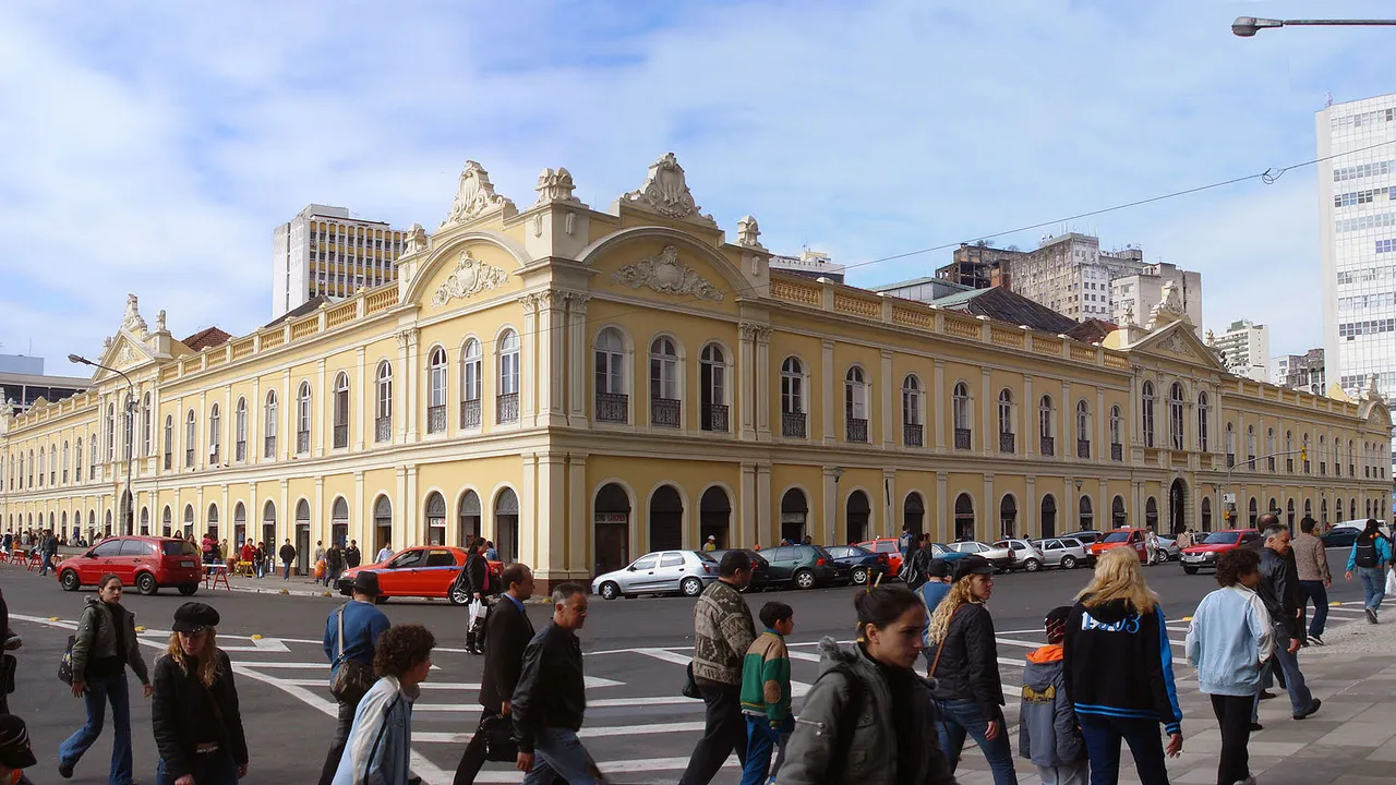 Fachada de um casarão eclético amarelo de dois andares, com pátio movimentado de gente e carros em primeiro plano e prédios modernos ao fundo — o Mercado Público de Porto Alegre.