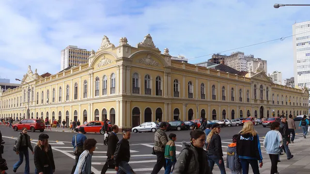 Fachada de um casarão eclético amarelo de dois andares, com pátio movimentado de gente e carros em primeiro plano e prédios modernos ao fundo — o Mercado Público de Porto Alegre.