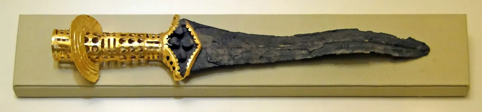 Punhal de bronze com cabo de folha de ouro recortada e vazada, encontrado em Malia, Creta, datado de 1800–1700 a.C., exposto sobre uma base clara no Museu Arqueológico de Heraclião