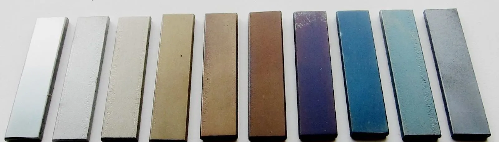Dez barrinhas de aço lado a lado sobre fundo claro: as duas primeiras em cinza, e as seguintes em tons crescentes de palha, dourado, marrom, púrpura, azul e cinza-azulado.