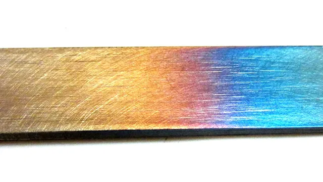 Barra chata de aço, em close, com a superfície mudando de cor ao longo do comprimento: palha e dourado de um lado, passando por marrom e púrpura até azul do outro — as cores de revenido.