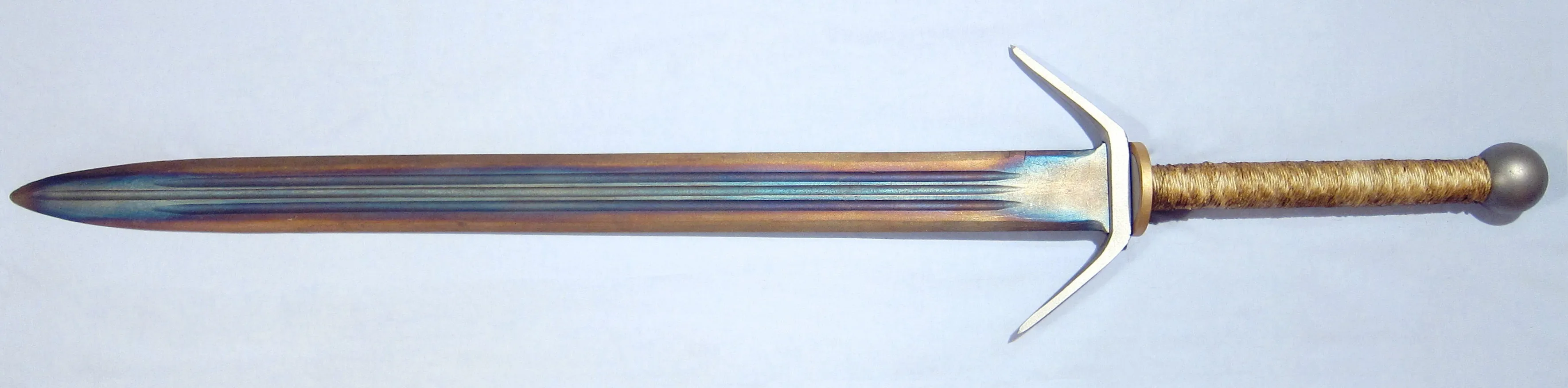 Espada de lâmina reta sobre fundo claro, com a lâmina mostrando faixas de cor: dourado perto do fio e azul e púrpura ao longo do centro e da guarda; cabo enrolado em cordão e pomo redondo.