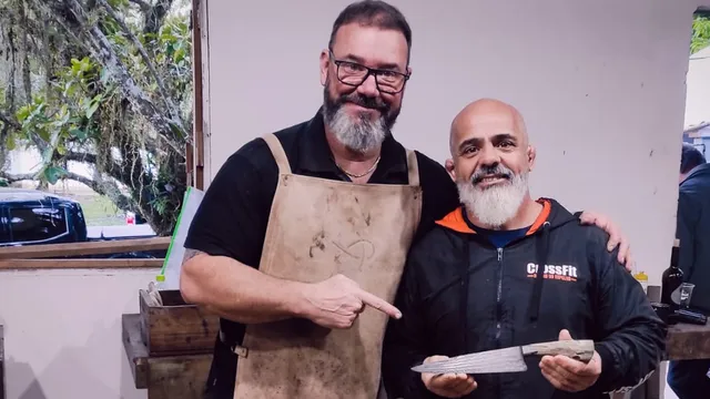 Sandro Boeck, de avental de couro, aponta sorrindo para Walter Fonseca, que segura com as duas mãos uma faca de damasco com cabo claro, dentro da oficina.