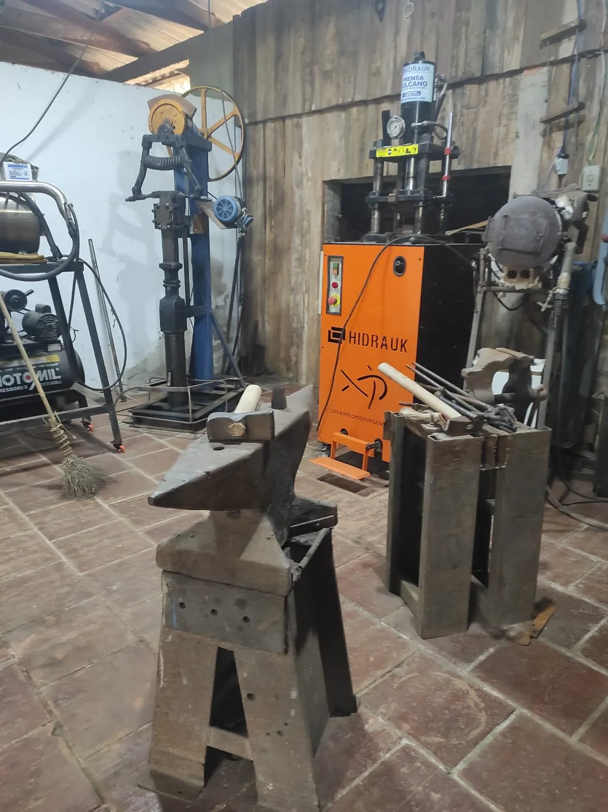 Interior do galpão da Peixinho Moreno Cutelaria: bigorna sobre cavalete de ferro, martelete pneumático azul ao fundo, prensa hidráulica Hidrauk laranja com a marca da casa e compressor à esquerda.